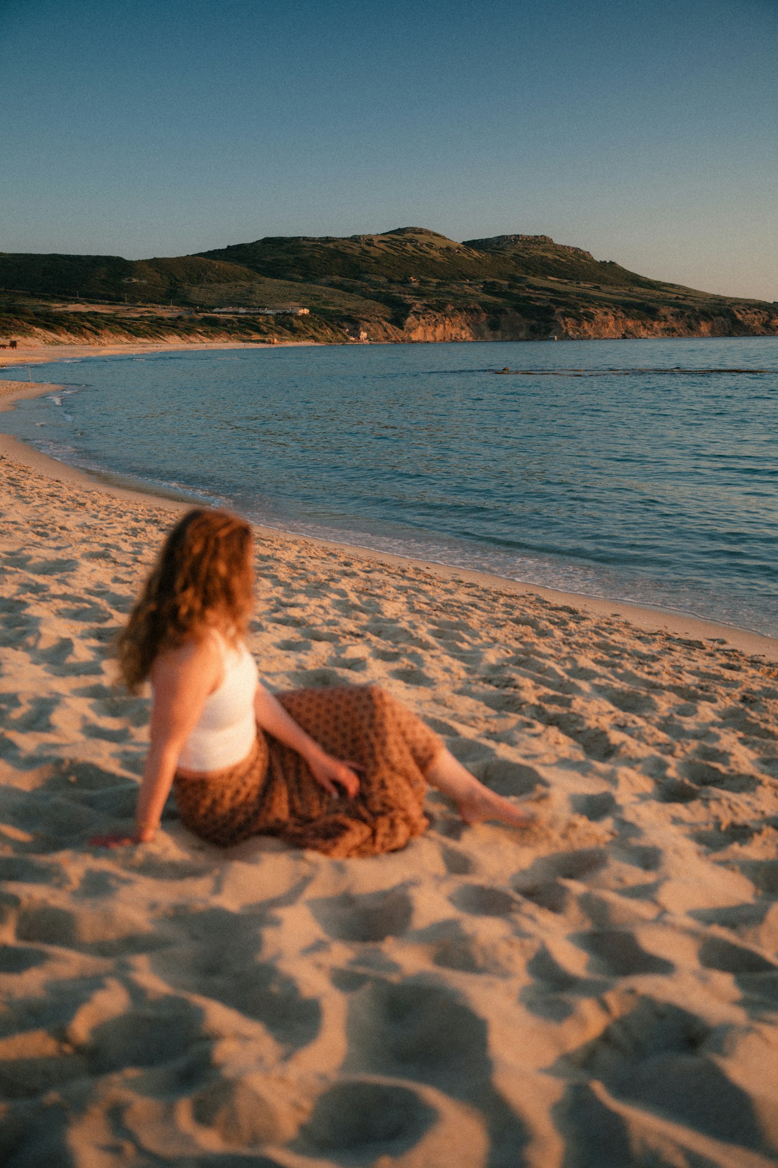 A woman sitting on the sand, looking out toward the sea at golden hour